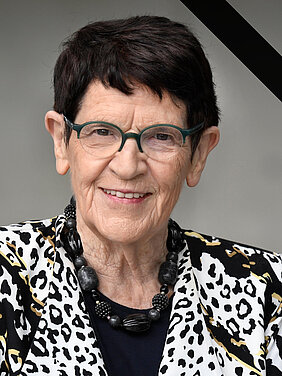 Rita Süssmuth vor neutralem Hintergrund. Sie schaut freundlich in die Kamera, trägt ein gemustertes Jackett. Das Bild ist oben rechts mit einer schwarzen Binde als Zeichen der Trauer versehen.